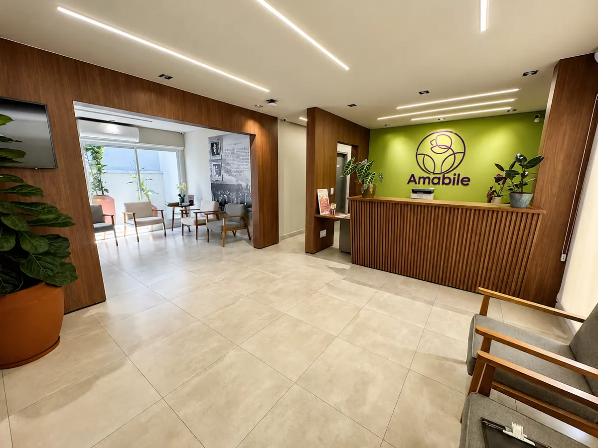Recepção da Clínica Amabile: balcão de madeira ripada, parede verde com a logo Amabile, plantas e sala de espera iluminada ao fundo